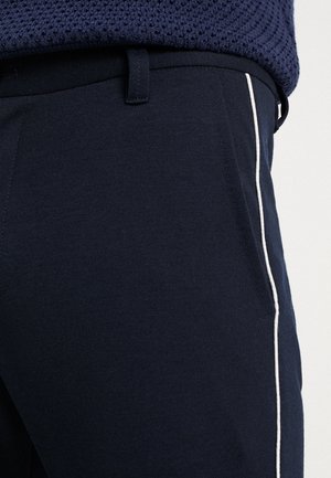 Granatowe spodnie z białym paskiem po bokach, noszone z fakturowanym granatowym topem, pokazujące szlufki na pasek i kieszeń po prawej stronie.