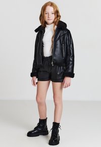 Giacca di pelle nera con colletto in pelliccia sintetica, pantaloni corti di pelle nera e stivali neri con lacci e suole spesse. La modella ha lunghi capelli rossi ondulati.