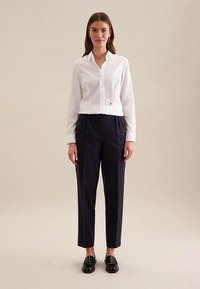 Biała skrócona koszula na guziki z długimi rękawami, zestawiona z granatowymi plisowanymi spodniami i czarnymi mokasynami. Gładka tkanina, dopasowany krój.