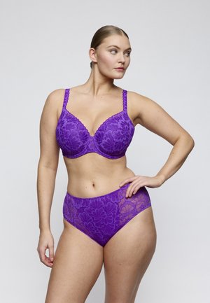 Vrouw poseert in paarse kanten beha en hoge taille slip, kijkt opzij tegen een effen lichte achtergrond.
