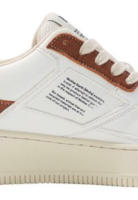 Witte synthetische sneakers met bruine leren accenten, een gestructureerde beige tong en oogjes. Een tekstlabel aan de zijkant geeft details over duurzame kenmerken.