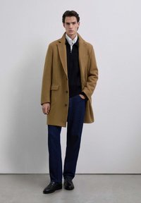 Joven de pie contra una pared blanca, vistiendo un abrigo color camel, chaqueta negra con cremallera, camisa blanca, pantalones azules y mocasines negros.