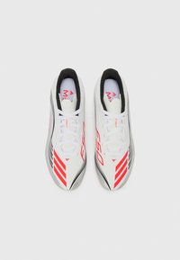 Crampons de football blancs avec des accents rouges et noirs, présentant un design épuré, une texture lisse et des rayures marquées sur les côtés.
