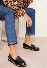 Mocasines de charol negros con un acabado suave, presentan una tira decorativa en la parte frontal, combinados con jeans de mezclilla azules cortos.