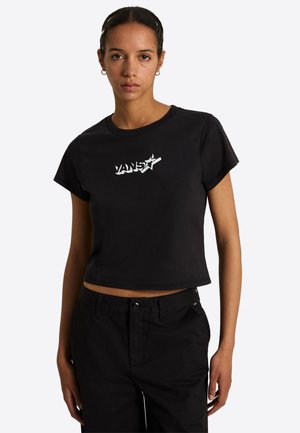 Jeune femme portant un t-shirt court noir Vans avec logo blanc et un pantalon noir, debout devant un fond clair uni.
