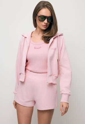 Vrouw draagt een roze hoodie met ritssluiting, bijpassende korte broek, roze hemd en donkere zonnebril, poserend tegen een effen lichte achtergrond.