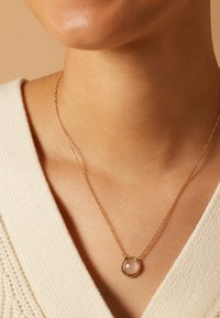 Gouden ketting met een ronde rozenkwarts hanger, geplaatst in een onregelmatig gevormde houder. Geweven stoffen achtergrond, neutrale kleur.