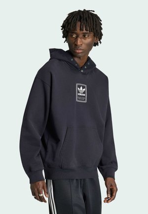 Homme portant un sweat à capuche noir Adidas avec logo à l'avant et un pantalon noir Adidas avec des bandes blanches, regardant légèrement vers sa gauche.
