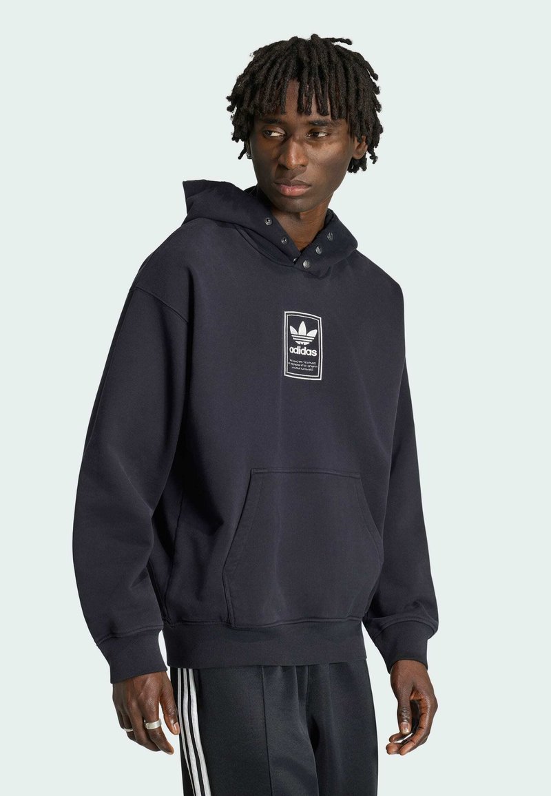 Homme portant un sweat à capuche noir Adidas avec logo à l'avant et un pantalon noir Adidas avec des bandes blanches, regardant légèrement vers sa gauche.