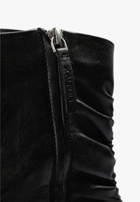 Botín de cuero negro con diseño fruncido, textura brillante y cremallera metálica plateada en el lateral para cierre.