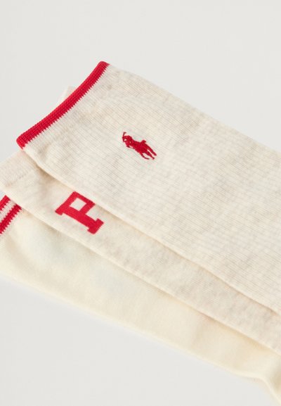 Ponožky po kolena v krémové bavlně s červenými ribbed manžetami. Obsahují malé červené logo a výrazný text blízko vrchu. Hladká textura, lehké.