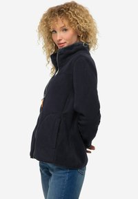 Marininspirerad fleecejacka med hög krage, dragkedja framtill, två sidofickor och strukturerad tyg. Modellen står åt sidan, iförd jeans.