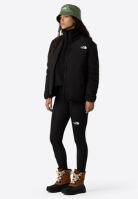 Chaqueta aislante negra, leggins negros, botas marrones y negras con cordones blancos, y un sombrero tipo cubo verde oliva. Logos en la chaqueta y los leggins.