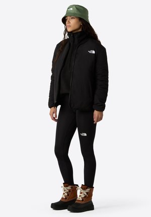 Sort isoleret jakke, sorte leggings, brune og sorte støvler med hvide snørebånd, og en olivengrøn bøttehat. Logoer på jakken og leggingsene.