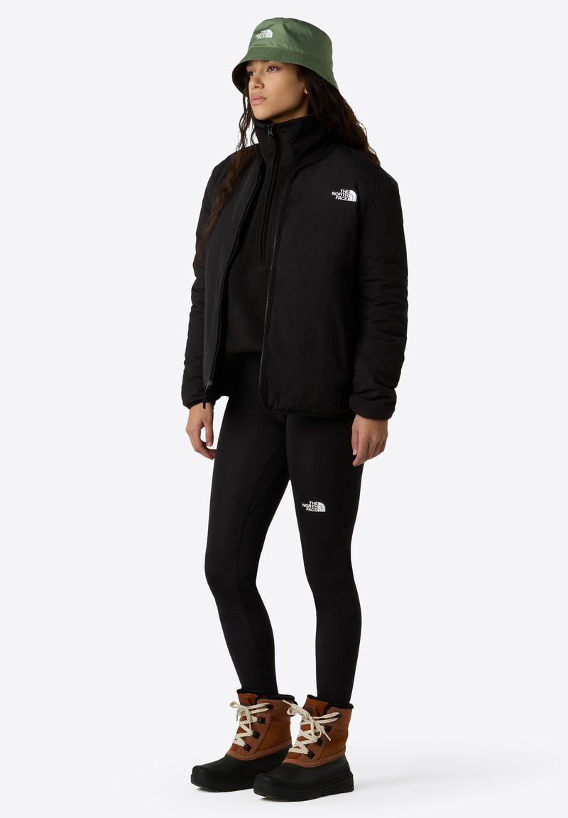 Chaqueta aislante negra, leggins negros, botas marrones y negras con cordones blancos, y un sombrero tipo cubo verde oliva. Logos en la chaqueta y los leggins.
