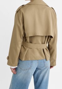 Person wearing a beige belted trench coat and light blue denim jeans, shown from the back against a white background.