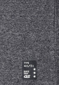 Tumma harmaa teksturoitu kangas mustalla tarralla. Tarrassa on "TYPE 40/51" ja "SINCE 1994 QS" valkoisella kirjotettuna ja kolme tähteä.