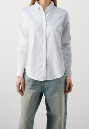 Vit skjorta med knäppning framtill, långa ärmar, krage och rundad nederkant, gjord av en mjuk tyg; kombinerad med ljusblå denimjeans.