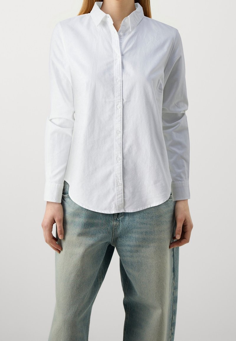 Camicia bianca con bottoni, maniche lunghe, colletto e orlo curvo, realizzata in un tessuto morbido; abbinata a jeans in denim azzurro chiaro.