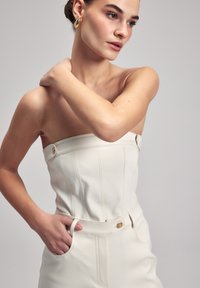 Jumpsuit bianco in pelle senza spalline con una silhouette aderente, cuciture verticali, tasche laterali, dettagli con bottoni dorati e una texture liscia.