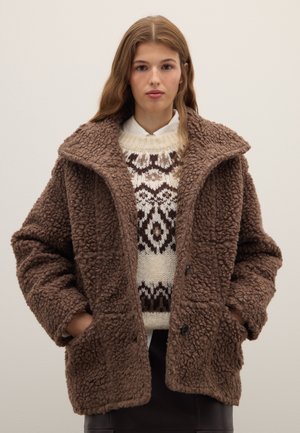 Chaqueta marrón y peluda con acabado texturizado, gran cuello, bolsillos laterales y botones negros, llevada sobre un suéter de punto con estampado.