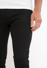 Černé džínové kalhoty s úzkým střihem, s hladkou texturou a standardním pětiprázdnovým designem, částečně zobrazené s rukou opřenou o bok.