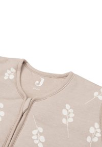 Combinaison bébé zippée beige clair avec un col rond, ornée de motifs de feuilles blanches et d'une étiquette avec "J" et la taille "90 cm" à l'intérieur.