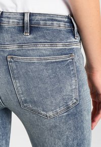Jeans de denim azul deslavado con un ajuste de talle medio, costuras de bolsillo visibles y un diseño clásico de bolsillo trasero. La textura parece desgastada y suave.