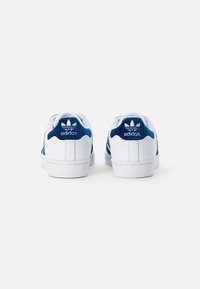 White sneakers with blue accents, featuring a smooth leather upper and textured suede heel. Adidas logo and three stripes design visible.