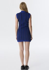 Gebreide jurk in blauw met een zwart geometrisch patroon, mouwloos ontwerp, hoge kraag en geribde zoom. Effen zwarte schoenen maken de look compleet.