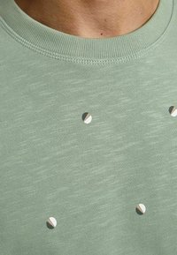 Jasnozielona bluza z okrągłym dekoltem i teksturowaną tkaniną, ozdobiona małymi, kolorowymi kropkami w kolorze białym, brązowym i beżowym.