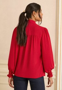 Crvena bluzica dugih, napuhanih rukava, s nabranoj manžetom i meko plisiranom pozadinom. Glatka tkanina, opušteni kroj i klasični dizajn ovratnika.