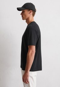 Schwarzes Baumwoll-T-Shirt mit kurzen Ärmeln, einem runden Ausschnitt und einer lockeren Passform, kombiniert mit einer schwarzen Mütze, vor einem hellen Hintergrund dargestellt.