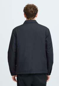 Man with curly hair wearing a black jacket, shown from the back against a plain light background.