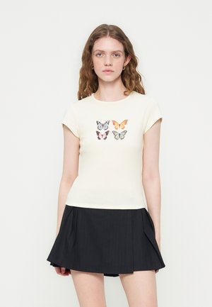 Νέα γυναίκα με κυματιστά μαλλιά φοράει κρεμ κοντομάνικη μπλούζα με τέσσερα σχέδια πεταλούδας και μαύρη πλισέ μίνι φούστα, στέκεται σε λευκό φόντο.