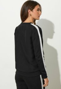 Donna con capelli castani che indossa una felpa nera e pantaloni con strisce bianche lungo le maniche e le gambe dei pantaloni, in piedi di lato contro un muro bianco.