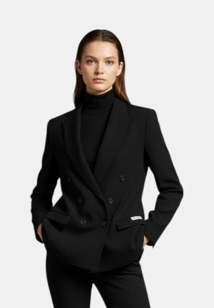 Donna con blazer doppiopetto nero e dolcevita nero, in piedi con le mani in tasca contro uno sfondo chiaro e uniforme.