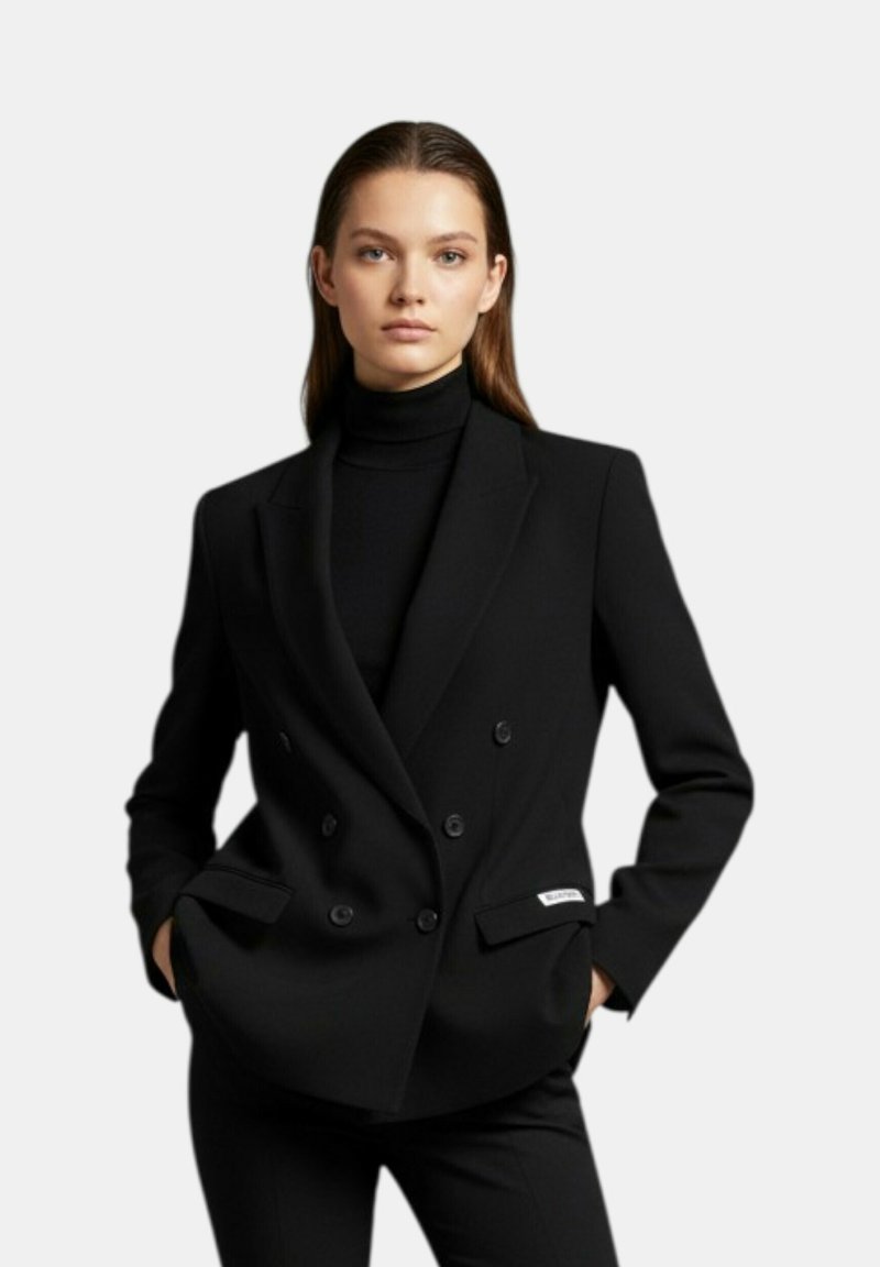 Donna con blazer doppiopetto nero e dolcevita nero, in piedi con le mani in tasca contro uno sfondo chiaro e uniforme.