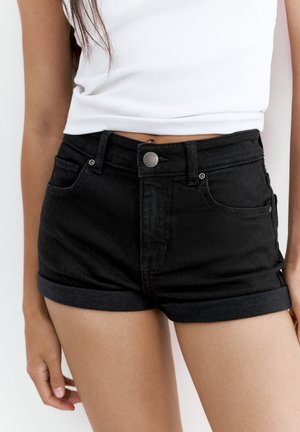 Kvinde iført rullede sorte denimshorts og en hvid ærmeløs top, stående med armene afslappet ned langs siden.
