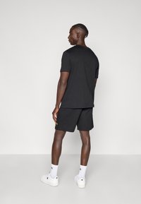 Μαύρο μπλουζάκι με κοντά μανίκια και στρογγυλή λαιμόκοψη, σε συνδυασμό με μαύρο σορτς. Φωτεινή κίτρινη λεπτομέρεια λογότυπου στο σορτς, φορεμένο με λευκά αθλητικά παπούτσια.