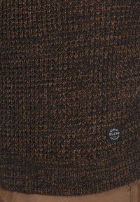 Brauner gestrickter Pullover mit einem strukturierten Muster und einem kleinen schwarzen Etikett mit weißem Text, der "DENIM BLEND with ATTITUDE" liest.