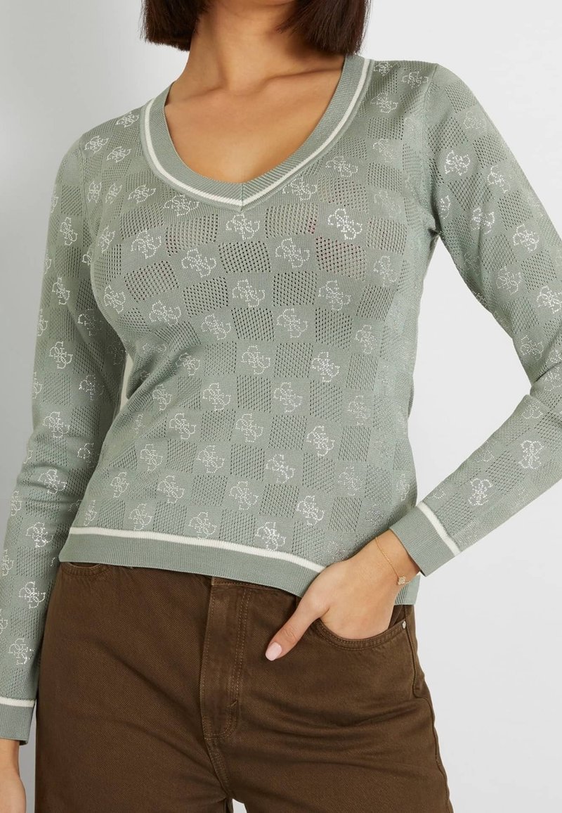 Femme portant un pull vert clair à motifs avec col en V et un pantalon marron, une main dans la poche, sur fond uni.