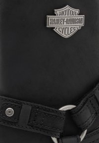 Botas de cuero negro con un logo metálico de Harley-Davidson, que cuentan con una correa con una hebilla circular plateada y un detallado cosido.