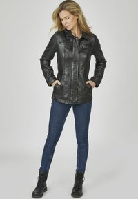 Schwarze Lederjacke mit tailliertem Schnitt, zwei Reißverschlusstaschen auf der Brust, Seitentaschen und einer glatten Oberfläche. Kombiniert mit dunklen Skinny-Jeans und schwarzen Stiefeln.