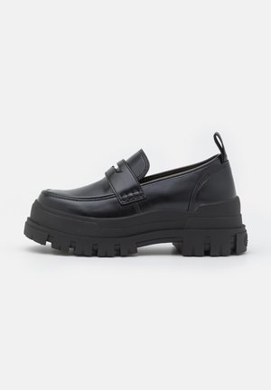 Sort læderloafer med tyk rillede sål, mok tå syning, remdetalje over toppen og trækstrop på hælen, vist i sideprofil.