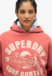 Sudadera en color frambuesa con un diseño estampado en blanco que muestra palmeras, un sol y un surfista. Capucha con cordón y tejido texturizado.