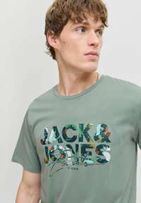 Zaļš kokvilnas T-krekls ar lielu, krāsainu "JACK & JONES Essentials" grafiku, apaļu kakla izgriezumu un īsām piedurknēm.
