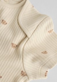 Țesătură bej deschis cu nervuri și modele mici cu ursuleți. Prezintă un guler rotund și mâneci scurte, textură moale.