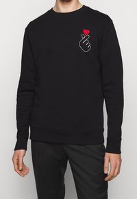 Sudadera negra de algodón, con un contorno blanco de una mano y un corazón rojo en el lado izquierdo. Puños y dobladillo acanalados.