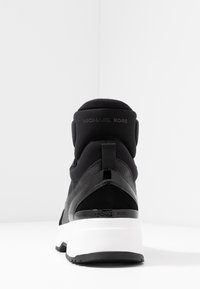 Černá teniska s texturovaným svrškem, hladkými akcenty a bílou podrážkou. Zadní část má tažnou smyčku a vypouklé logo.